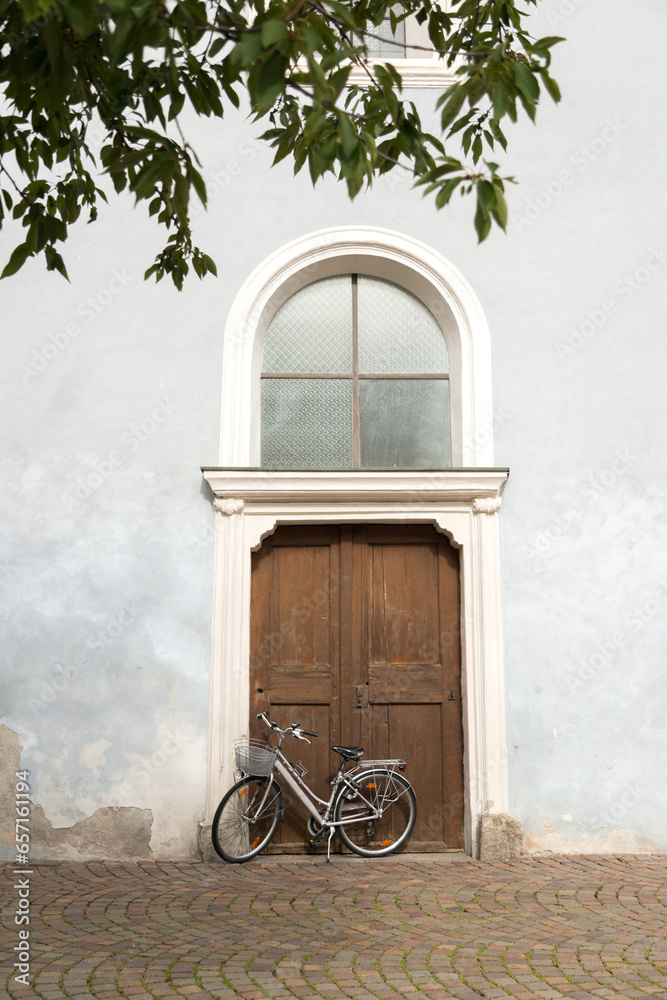 Postal de italia con portal y bicicleta apoyada sobre la fachada blanca con hojas verdes de árbol. Fondo vintage bonito de postal de Italia. Brixen. 