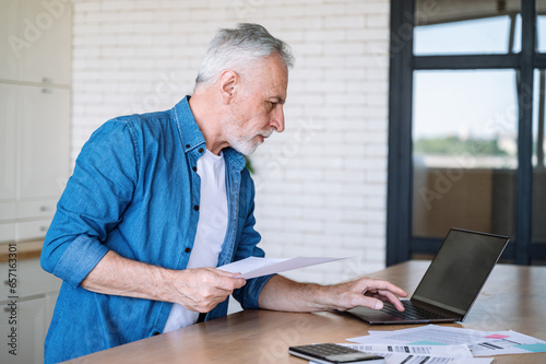 Middle aged man with paper document reviewing bank account on laptop