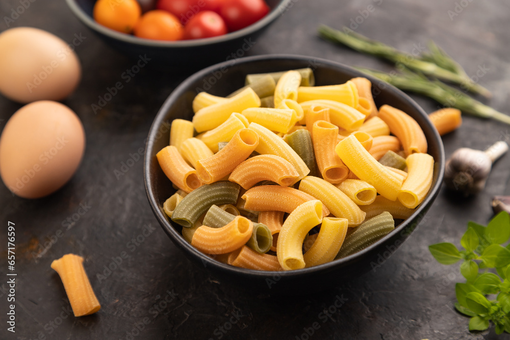 Rigatoni colored raw pasta with tomato, eggs, spices, herbs on black concrete. Side view, selective focus. Rigatoni colored raw pasta with tomato, eggs, spices, herbs on black concrete. Side view, selective focus.