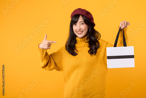 Joyful young Asian woman in her 30s, holding a shopping paper bag, pointing her finger, radiating happiness in a yellow sweater and red beret against a yellow background. photo