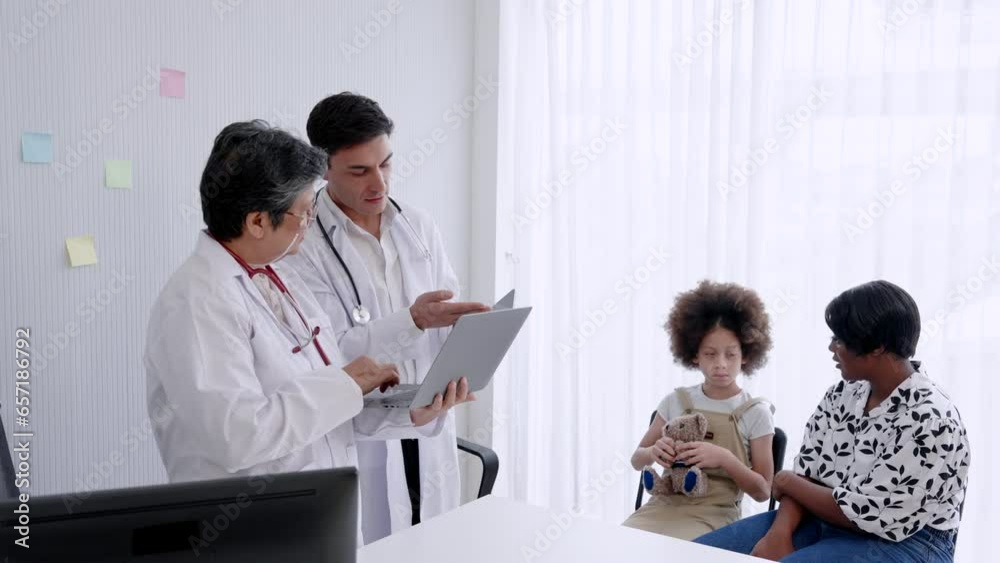Elderly Asian female doctor stands talks with male medical assistant, Information about patient condition is entered into system, In background is sick American girl mother brought child see doctor. Elderly Asian female doctor stands talks with male medical assistant, Information about patient condition is entered into system, In background is sick American girl mother brought child see doctor.
