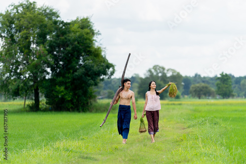 Young Asian girl and boy hold rice seedlings and plow walk together on road in rice field with big tree on the background.