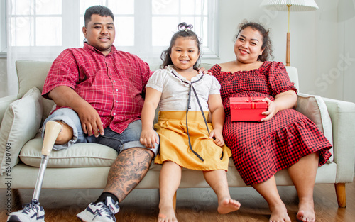 Portrait three people asian family, happy father, husband with disable prosthetic leg, giving red gift box to celebrate birthday, sitting on sofa at home, looking at camera. Healthcare Concept.