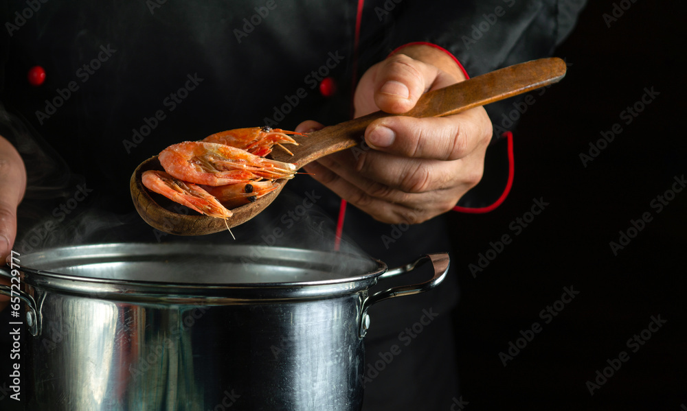 Professional chef prepares delicious shrimp in a pan in the kitchen. The concept of a delicious shrimps breakfast for a hotel. Spoon in the cook hand wis scampi - obrazy, fototapety, plakaty 