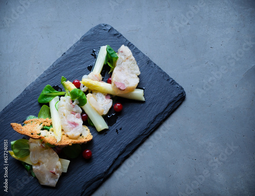 A gourmet seafood salad featuring white asparagus, mixed greens, red berries, and crispy bread, elegantly arranged on a black slate plate