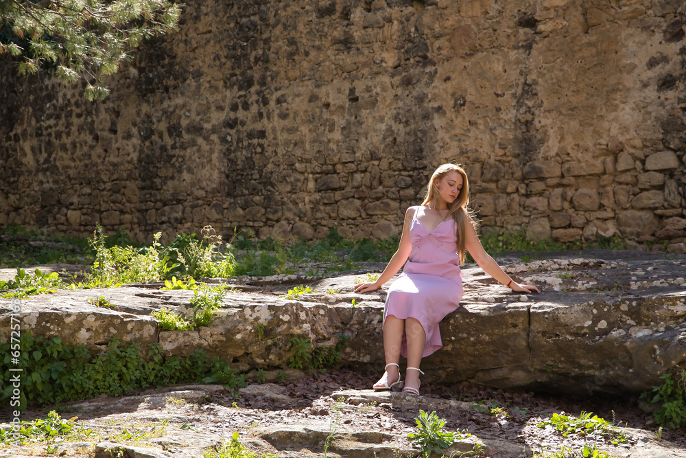 Young, beautiful, blonde woman in a pink dress, sitting on a stone inside an ancient medieval castle receiving the rays of the sun, alone, quiet and peaceful. Concept beauty, relax, tranquility, calm Young, beautiful, blonde woman in a pink dress, sitting on a stone inside an ancient medieval castle receiving the rays of the sun, alone, quiet and peaceful. Concept beauty, relax, tranquility, calm