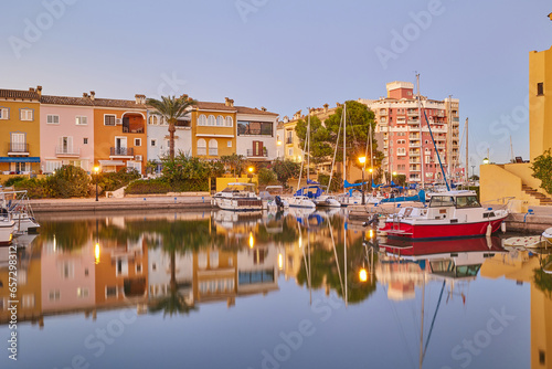 Panoramic view of colorful houses and moored yachts in the evening in Port Saplaya. Light from street lamps, buildings and boats reflects on the smooth surface of the water. Valencia's Little Venice.