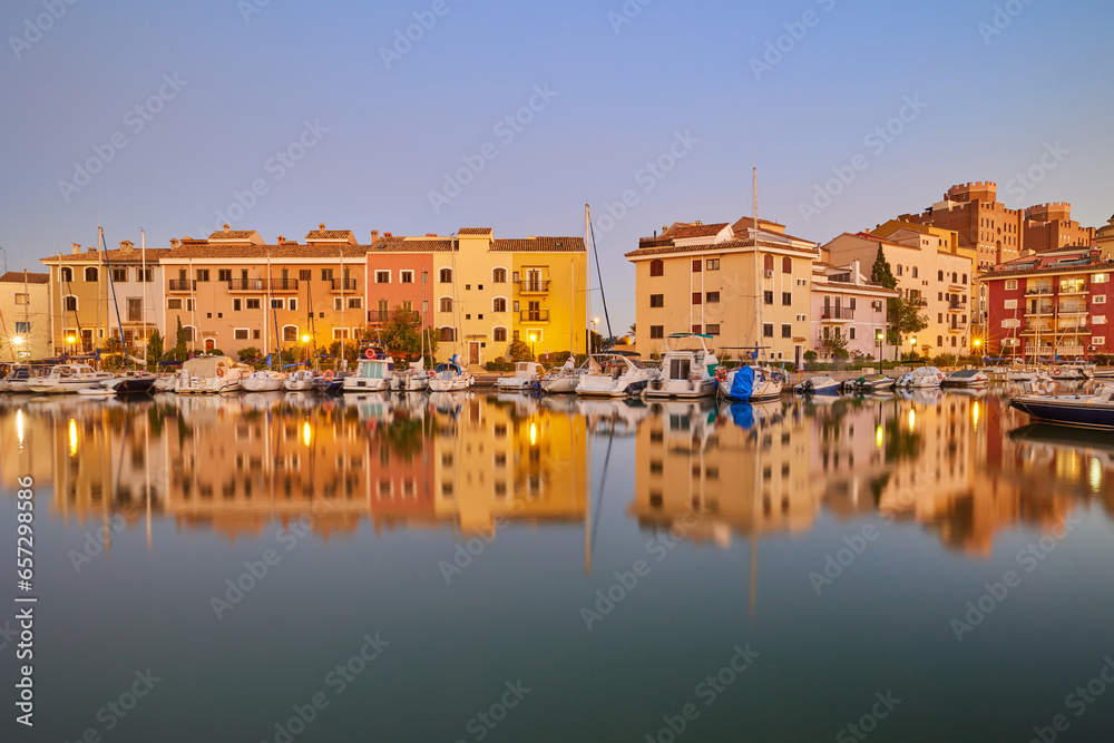 Panoramic view of colorful houses and moored yachts in the evening in Port Saplaya. Light from street lamps, buildings and boats reflects on the smooth surface of the water. Valencia's Little Venice.