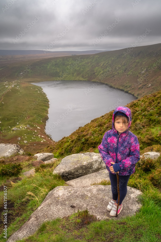 Young girl standing on the rock on the top of the mountain with lake, Lough Bray, down in background. Family hiking in Wicklow Mountains, Ireland