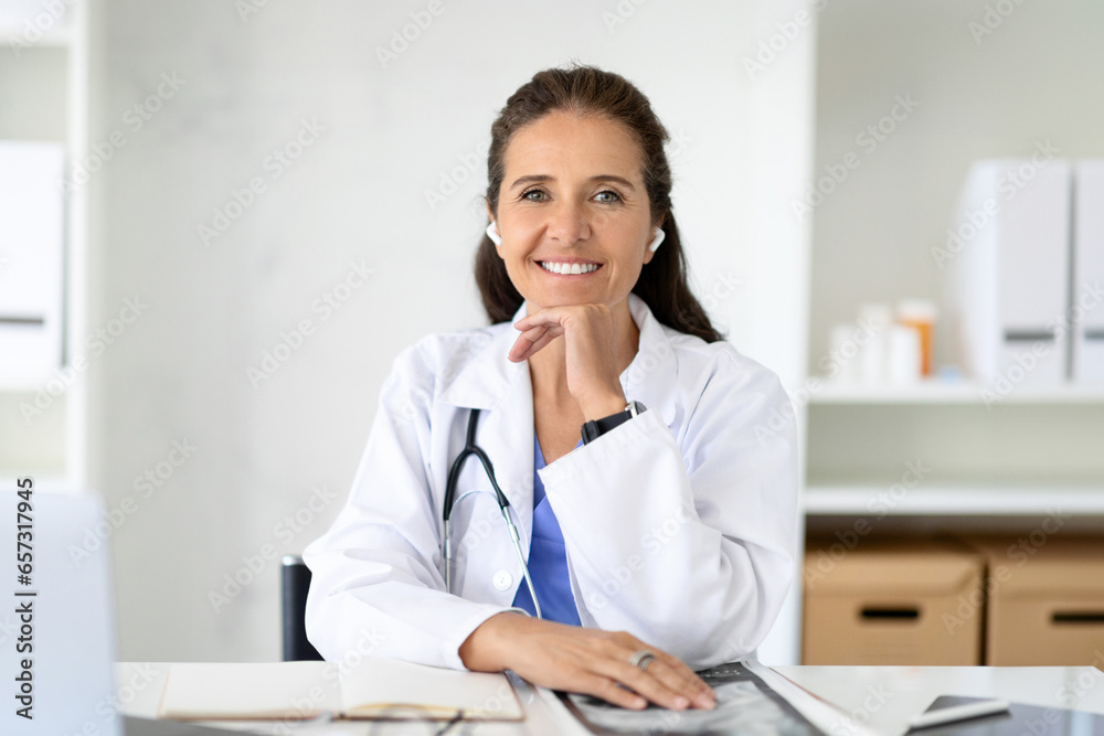 Cheerful middle aged woman doctor using wireless earphones. E-medicine, telehealth Cheerful middle aged woman doctor using wireless earphones. E-medicine, telehealth