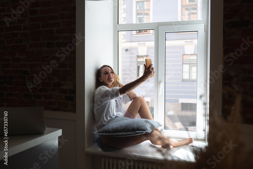 young beautiful woman talking on the phone sitting by the big window in the apartment