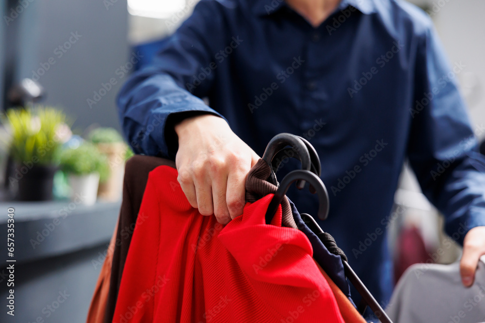 Close up of male hand holding clothes on hangers while shopping in clothing store, selective focus. Man customer choosing apparel at fashion outlet, looking though garment racks with clothing items - obrazy, fototapety, plakaty 