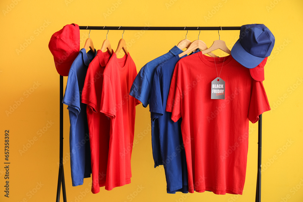 Obraz premium Rack with different t-shirts and caps on yellow background. Black Friday sale