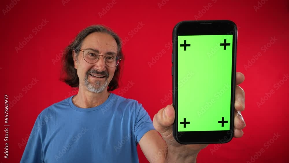 Excited mature elderly man puts hand on head and points to Big smartphone with blank green screen with tracking points, demonstrating copy space app or website design, mockup image on red background