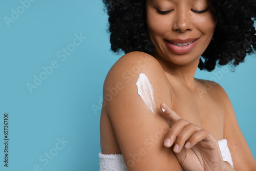 Young woman applying body cream onto shoulder on light blue background, closeup. Space for text