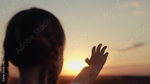 Cute little girl reaches hand to sunset sun disk making wish in evening field