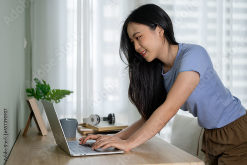 A beautiful Asian woman bending over a table and using her laptop, working in her room.