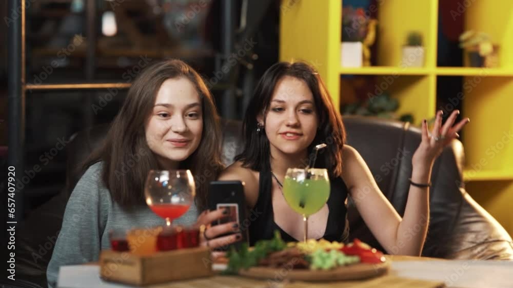 Two adult girls take selfies on smartphones while sitting in a bar with cocktails Two adult girls take selfies on smartphones while sitting in a bar with cocktails