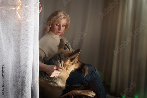 Adult mature woman of 40-60 with big shepherd dog in warm sweater. Room with calm cozy evening atmosphere with transparent curtains and soft warm light of lamps. Concept of love for animals and pets