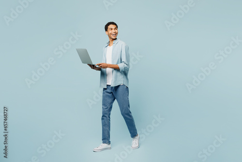 Full body side view young man of African American ethnicity wear shirt casual clothes hold use work on laptop pc computer look aside on area isolated on plain pastel light blue cyan background studio.