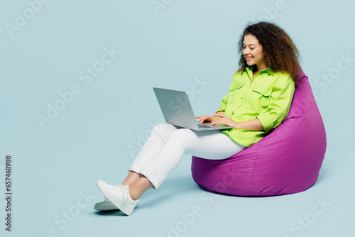 Full body young IT latin woman she wear green shirt casual clothes sit in bag chair hold use work on laptop pc computer isolated on plain pastel light blue cyan background studio. Lifestyle concept.