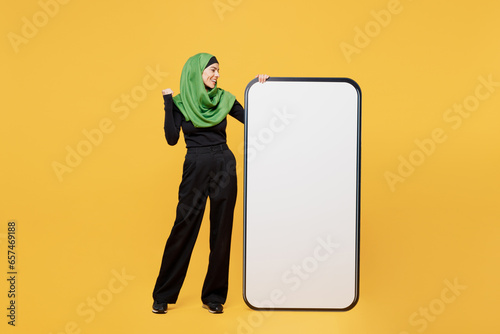 Full body young asian muslim woman in green hijab abaya black clothes big huge blank screen mobile cell phone do winner gesture isolated on plain yellow background People uae islam religious concept