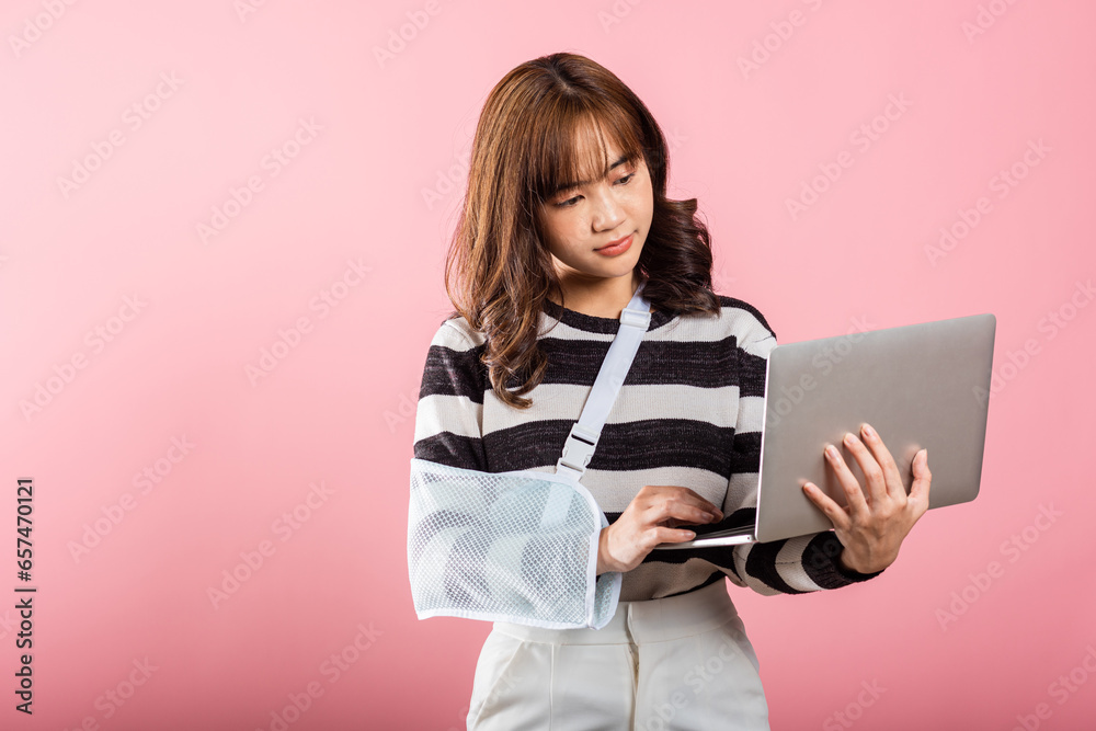 Fototapeta premium In a studio shot on a pink background, an Asian businesswoman showcases her confidence with a broken arm and arm splint. She continues her work on a laptop. Copy space included.