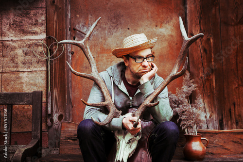 Pensive Man portrait in straw hat and glasses, old abandoned village background. man holds deer antlers in his hands. Adultery concept