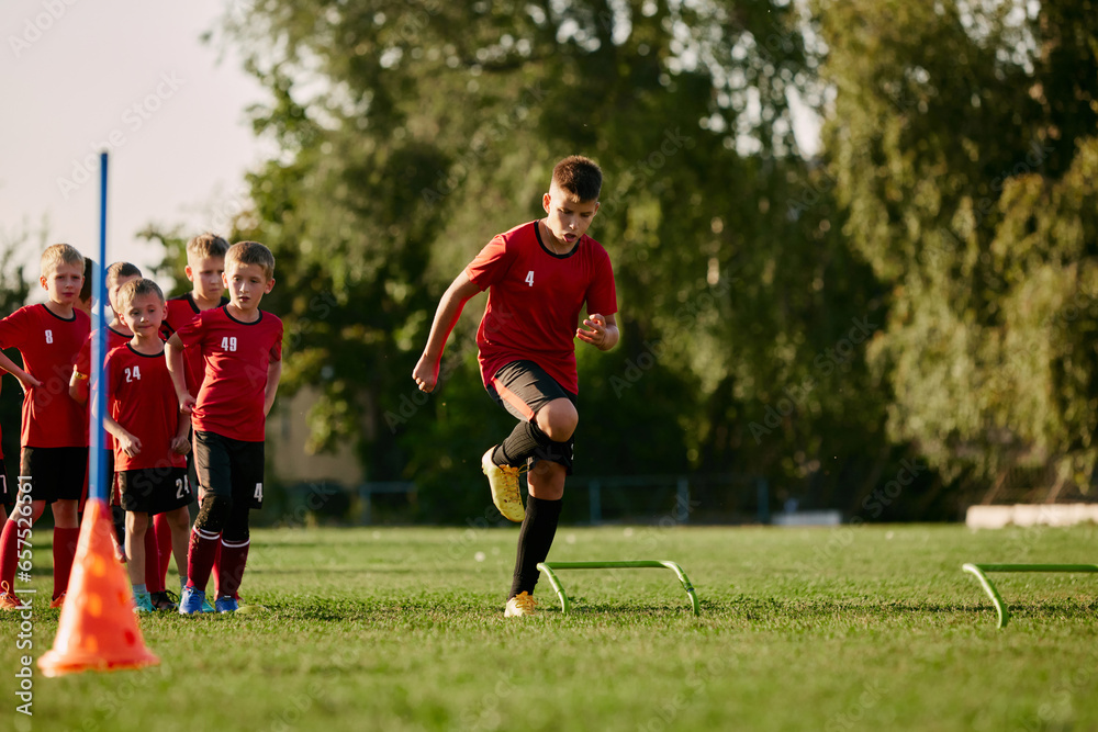 Full length of child, football player in sport uniform running, training, workout with football barriers in motion before match on soccer field. Full length of child, football player in sport uniform running, training, workout with football barriers in motion before match on soccer field.