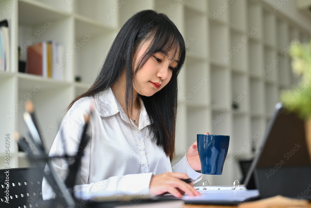 Asian female entrepreneur in white shirt drinking coffee and checking reports at desk Asian female entrepreneur in white shirt drinking coffee and checking reports at desk