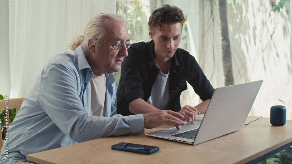 Medium shot of young grandson showing his grandfather how to use laptop while sitting at table at home Medium shot of young grandson showing his grandfather how to use laptop while sitting at table at home