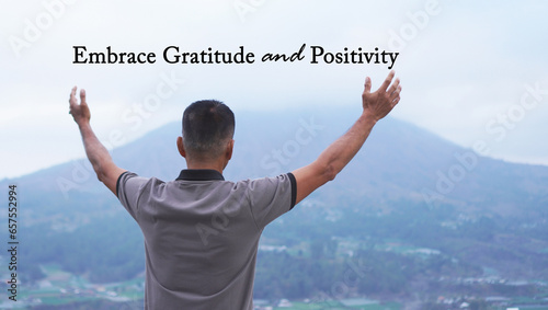 Inspirational quote - Embrace gratitude and positivity. With man standing alone from behind against the blue mountain view, raised hand and open arms. Gratefulness concept.