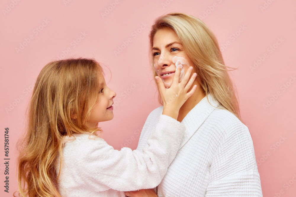 Evening hygiene. Smiling beautiful daughter, girl apply natural cosmetic cream to face, cheeks of young mother, woman over pastel pink background. Evening hygiene. Smiling beautiful daughter, girl apply natural cosmetic cream to face, cheeks of young mother, woman over pastel pink background.