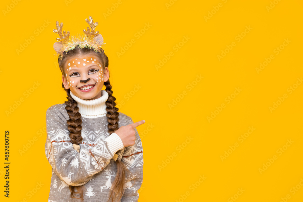A little girl in a Christmas sweater and with deer antlers points to your advertisement ad. Children's holiday makeup on the face. Copy space. Yellow isolated background. Space for text.