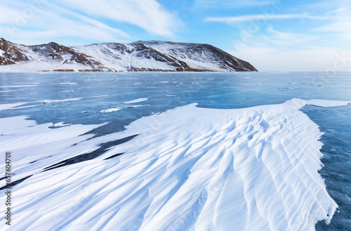 Winter landscape of frozen Baikal lake on cold February day. View of snow-capped coastal hills and iced bay with snow crusts in form of frozen waves form from wind on blue ice surface. Winter travel photo