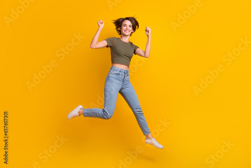 Full length photo of lucky pretty lady wear khaki t-shirt rising fists jumping high isolated yellow color background
