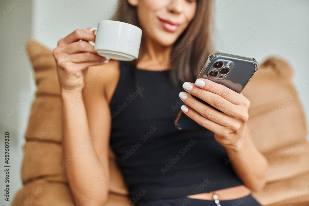 cropped view of happy woman using smartphone and holding cup of coffee at home, bean bag chair cropped view of happy woman using smartphone and holding cup of coffee at home, bean bag chair