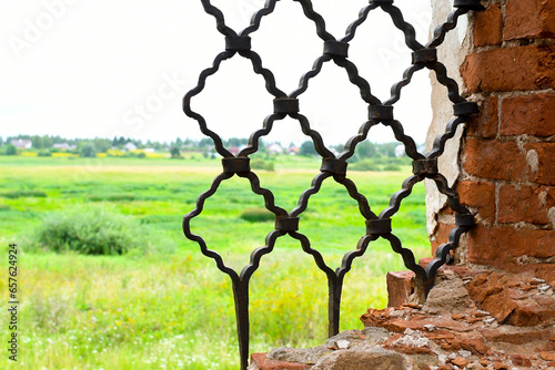 Wrought iron grille in the old window. An old dilapidated brick wall. From the window there is a view of a green field and a blue sky.