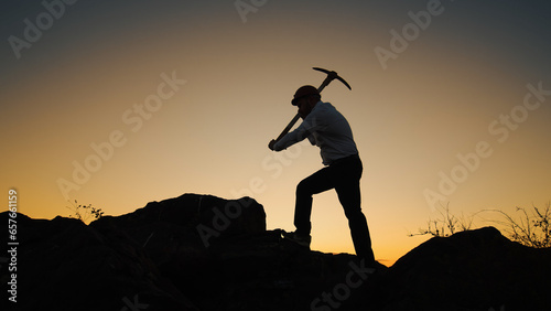 Fotografie Silhouette Man In Helmet With Pickaxe In His Hands Stands On Rock And Hits Stone