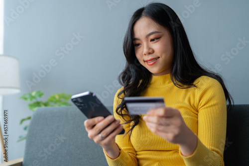 Young Asian woman holding credit card and smartphone sitting on sofa at home doing online banking transactions in home online shopping in living room. online payment and ordering concept.