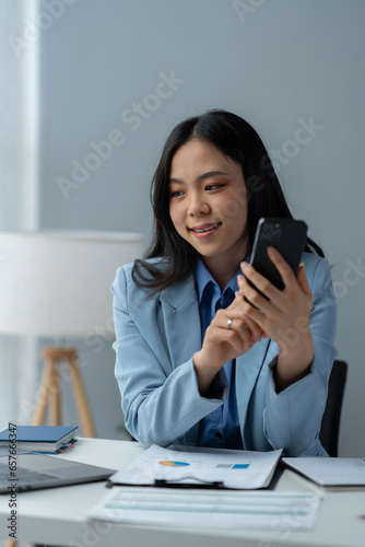 Young positive Asian female accountant using smartphone at her desk, contacting clients, checking business growth reports and relaxing on her mobile application. © crizzystudio