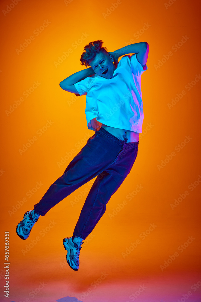 Full length portrait of young attractive man jumping put his hands behind head dressed casually against gradient orange background illuminated neon light.