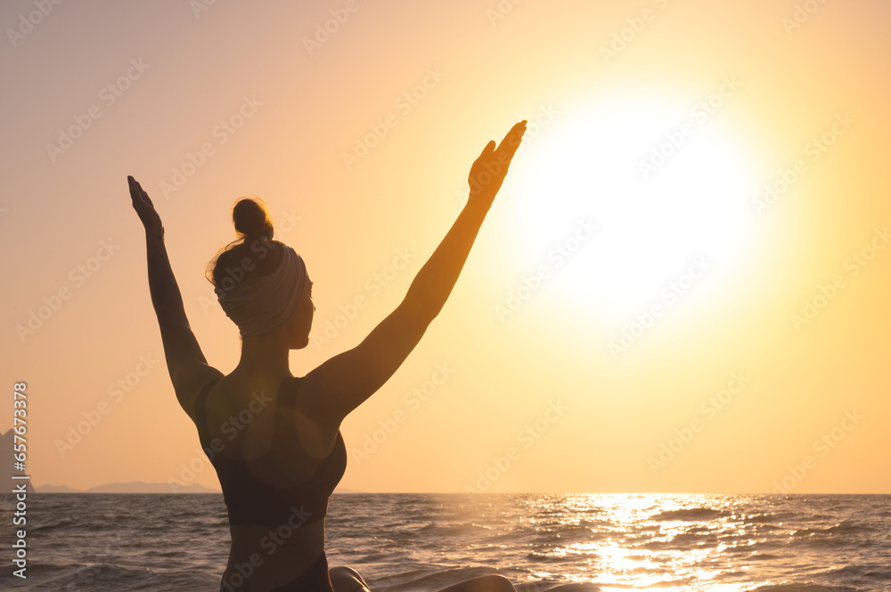 Fototapeta premium Free happy woman raises her hands and looks at the sun, rear side view. In the background, dawn, greeting the sun during yoga or meditation