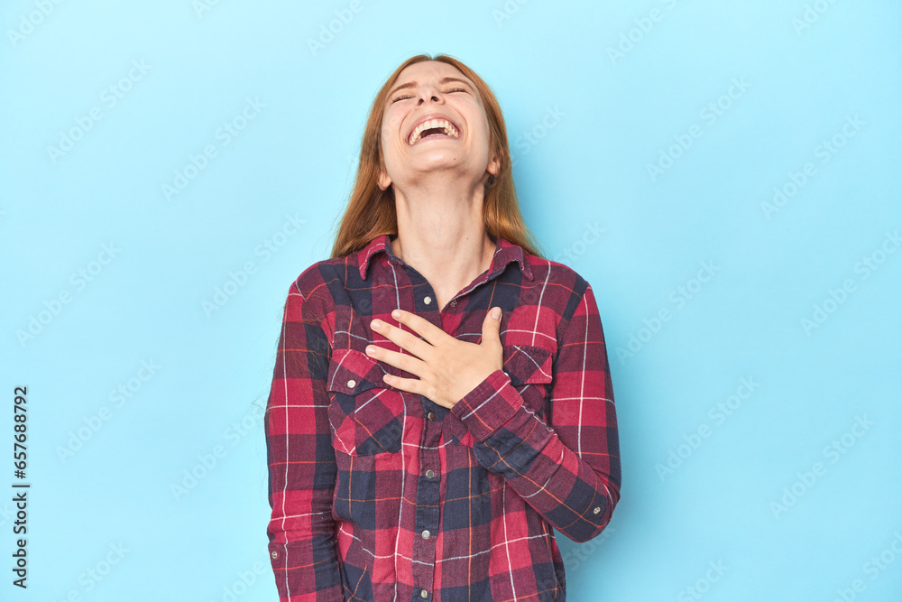 Redhead young woman on blue background laughs out loudly keeping hand on chest. Redhead young woman on blue background laughs out loudly keeping hand on chest.