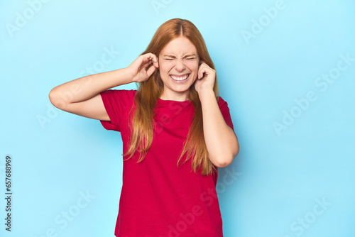 Redhead young woman on blue background covering ears with hands.