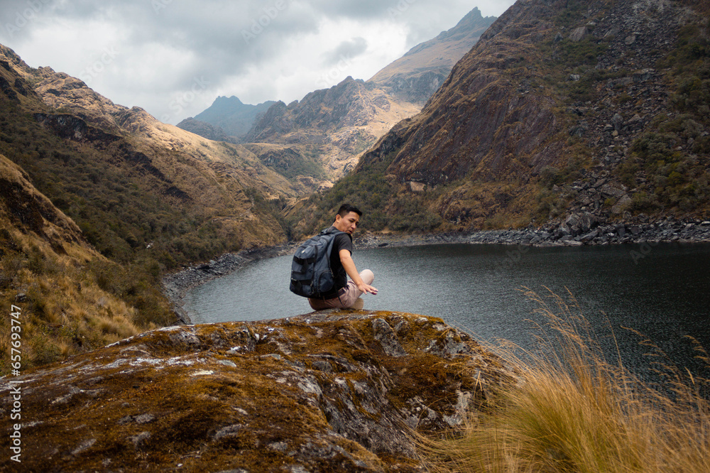 Un turista con mochila sentado con brazos abiertos en una roca a orillas de una laguna (Laguna Linda, Pachitea, Perú),paz,concepto de viajar,turismo Un turista con mochila sentado con brazos abiertos en una roca a orillas de una laguna (Laguna Linda, Pachitea, Perú),paz,concepto de viajar,turismo