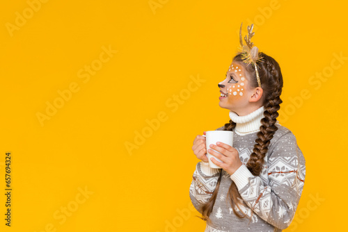 A child with deer antlers holds a cup of hot cocoa and looks at your advertisement. A little girl in a Christmas sweater. Yellow isolated background. Space for text. Copy space.
