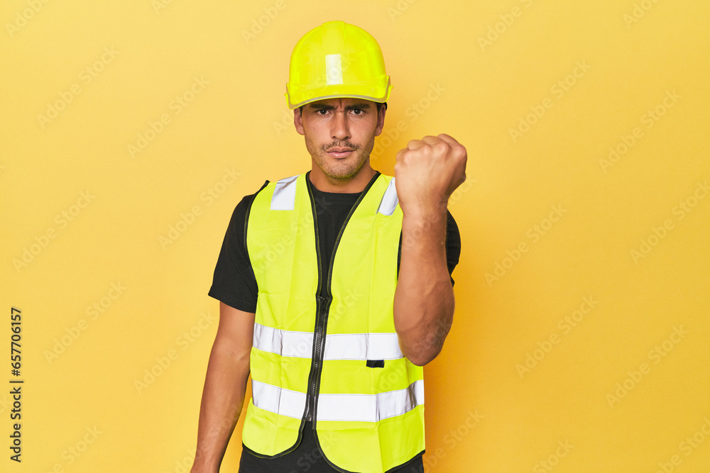 Latino worker in yellow vest and helmet showing fist to camera, aggressive facial expression. Latino worker in yellow vest and helmet showing fist to camera, aggressive facial expression.