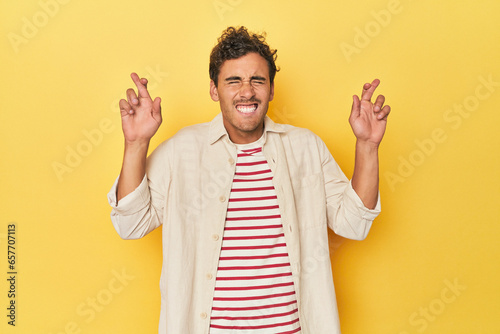 Young Latino man posing on yellow background crossing fingers for having luck