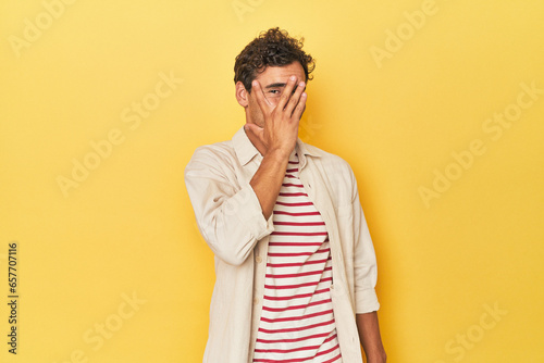 Young Latino man posing on yellow background blink at the camera through fingers, embarrassed covering face.